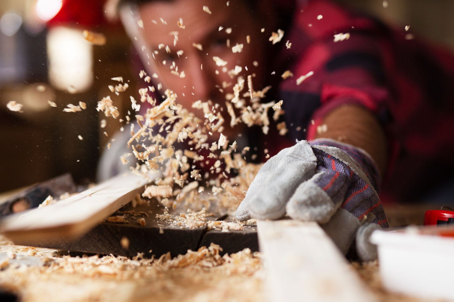 Ein Tischler bläst Späne von mehreren Holzbrettern nebeneinander und beurteilt sie dabei kritisch.
                Die Holzspäne fliegen.